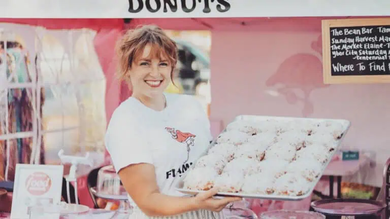 a baker holds up a plate of donuts in front of a tent at a local market