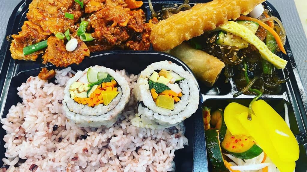 a hot meal to go featuring rice, sushi, fried spring rolls and veggies 