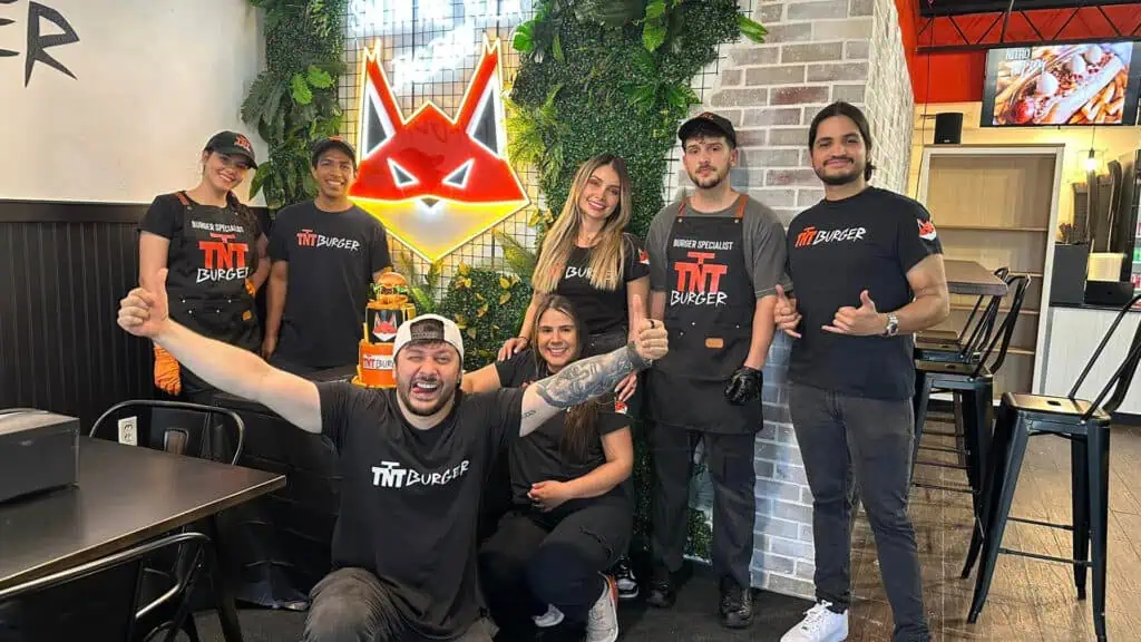 a group of people post in front of an ivy wall inside a burger restaurant. They're all wearing Black T Shirts celebrating the opening of a new restaurant