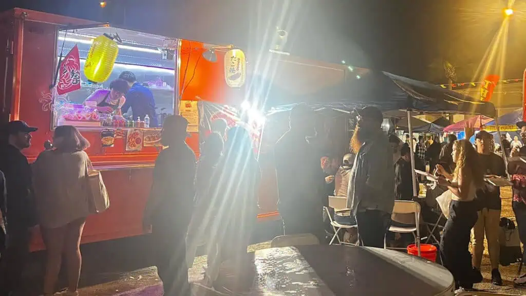 a food truck with asian lantern hanging over its hood. The vent is taking place at night and multiple people are waiting in line