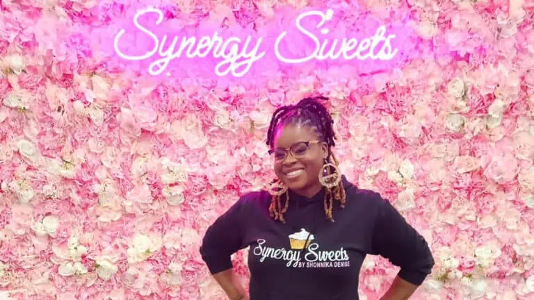 a perso poses in front of a flower wall with a pink neon sign that reads "Synergy Sweets"