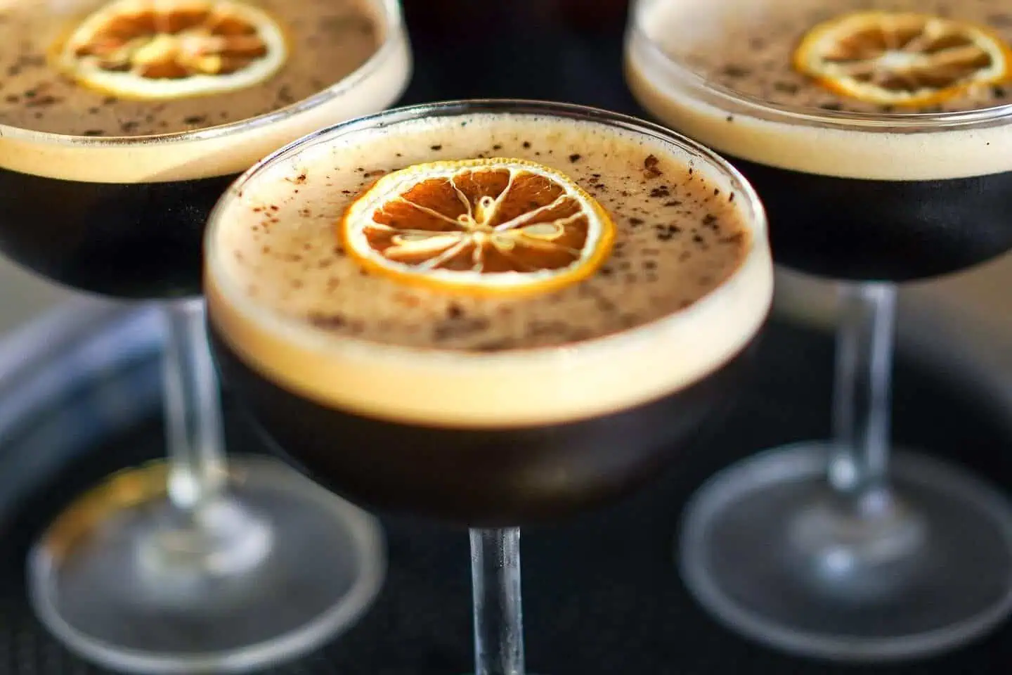 Three espresso martinis in cocktail glasses, each topped with a dried citrus slice and a layer of foam