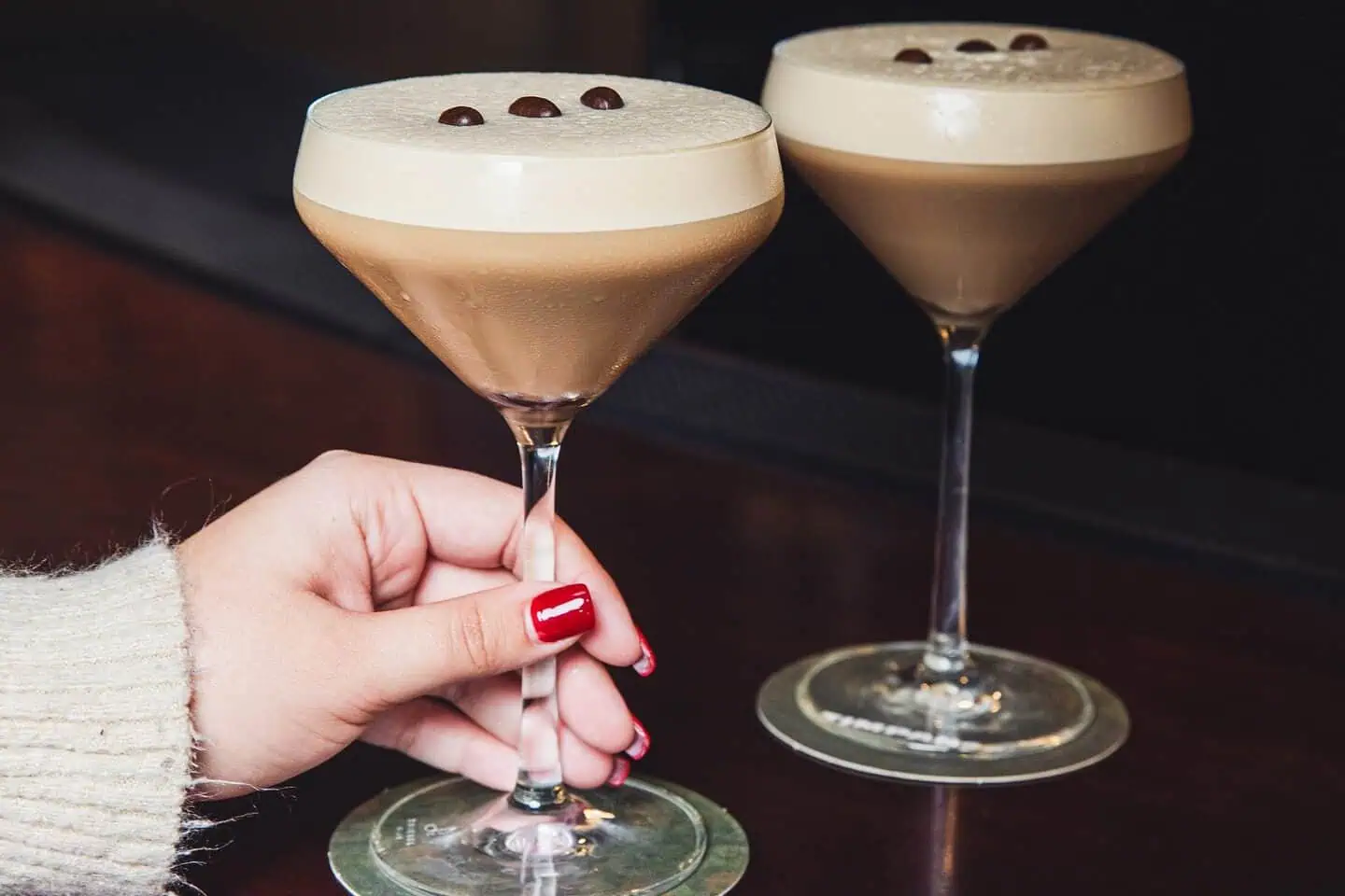 A hand with red nail polish holds a creamy espresso martini, with another identical drink in the background.