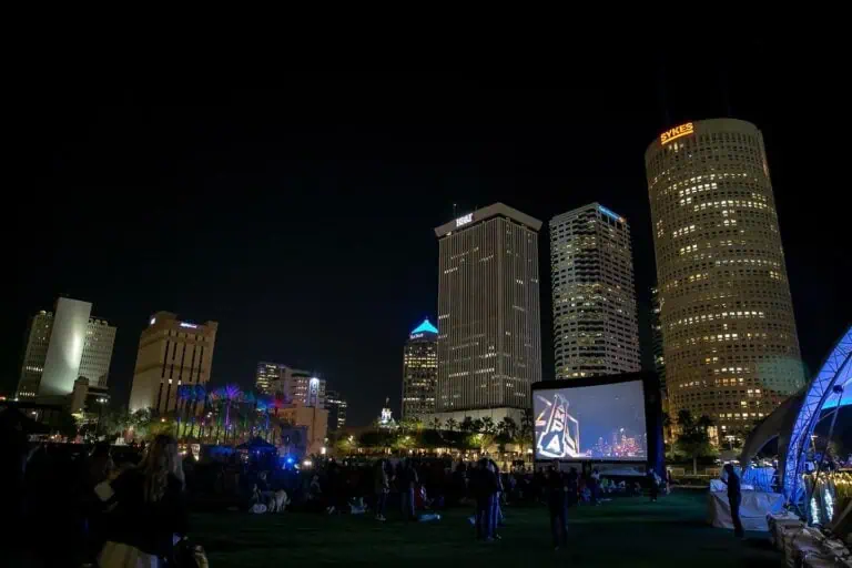 The Tampa skyline at night with a crowd gathered on a lawn for an outdoor movie screening.