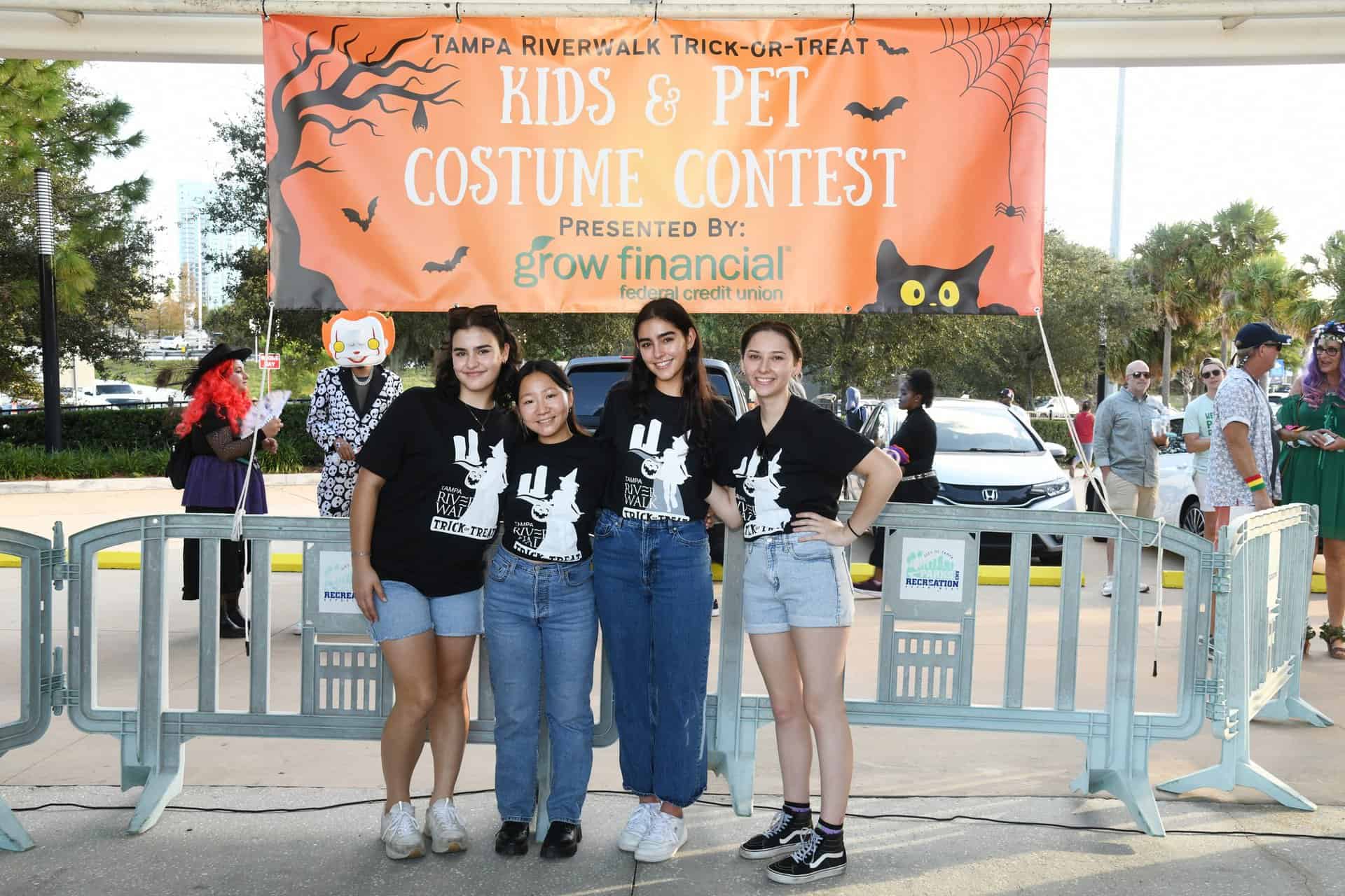 Four women in matching black shirts pose in front of an orange banner for a kids and pet costume contest at Tampa Riverwalk Trick-or-Treat.