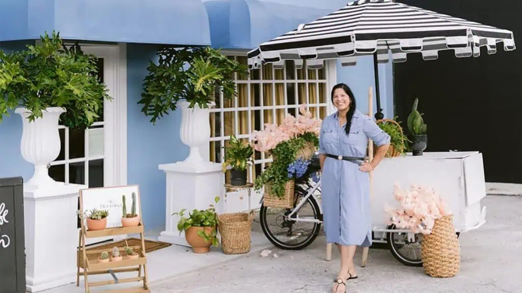 a person stands outside in a blue dress next to a small white cart with an umbrella opened.