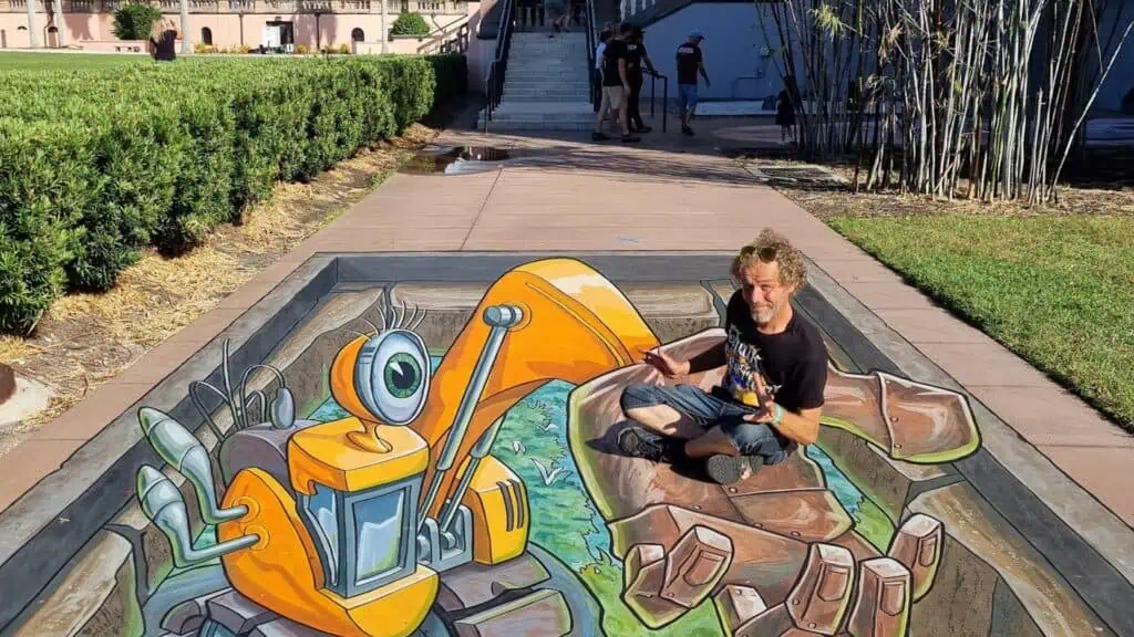 an artists poses with a 3D robot mural they painted on the street 