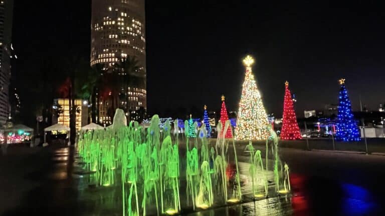 The Tampa Bay RiverWalk at night is decorated with colorful lit Christmas trees and illuminated fountains, with a tall building in the background.