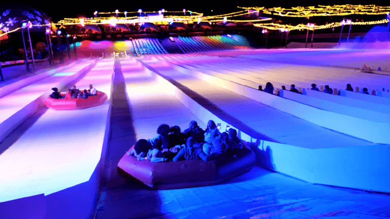 people on a tub going down a slop at night. the slope is lit up in purple and blue lights