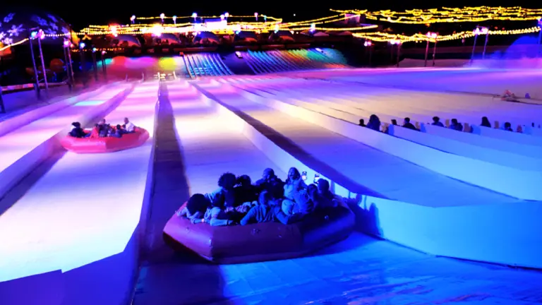 people on a tub going down a slop at night. the slope is lit up in purple and blue lights