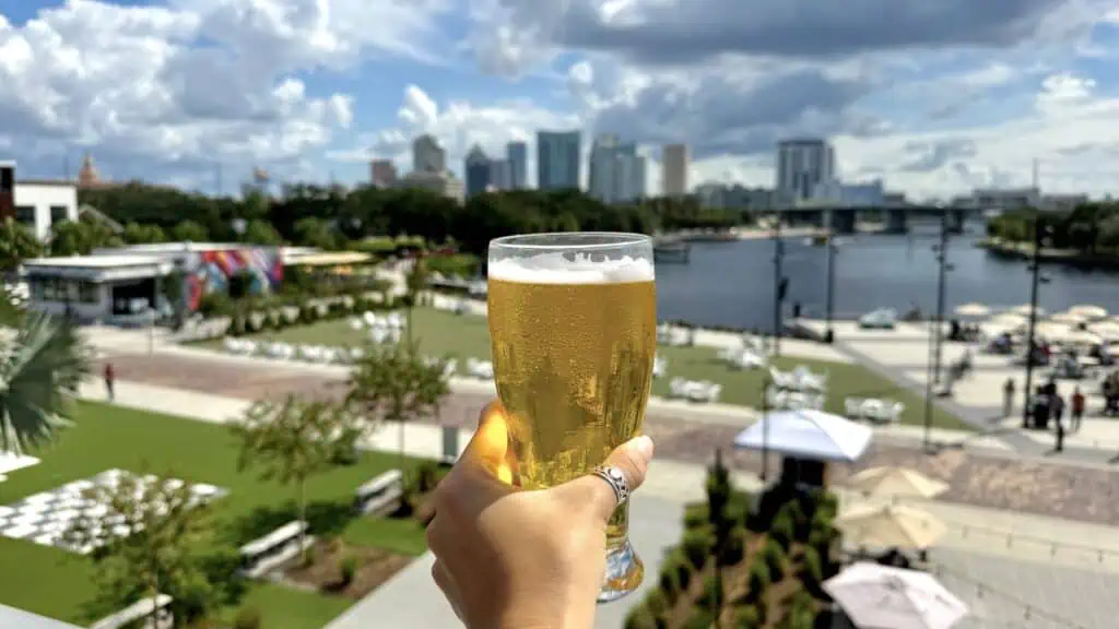 A hand holds a glass of beer against a scenic backdrop of a riverside park and the Tampa skyline.