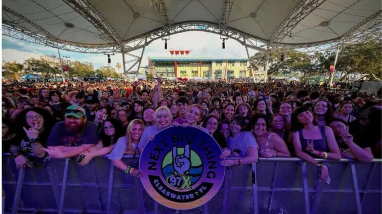 A lively crowd of fans gathers near the stage barricade under a large outdoor canopy at the "Next Big Thing" event hosted by 97X in Clearwater, Florida.