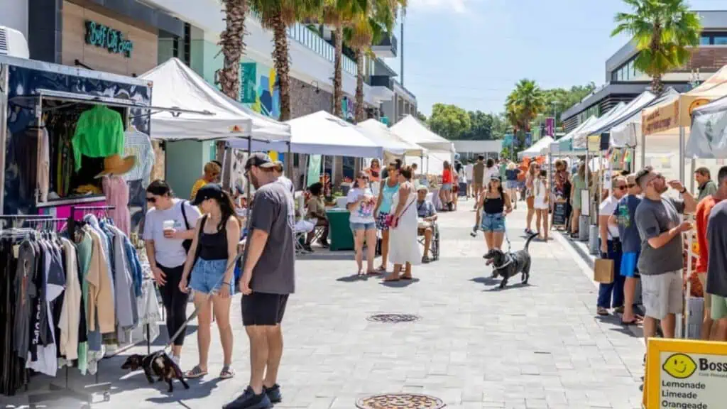 A bustling outdoor market with shoppers browsing stalls under white tents, surrounded by palm trees and bright sunlight.