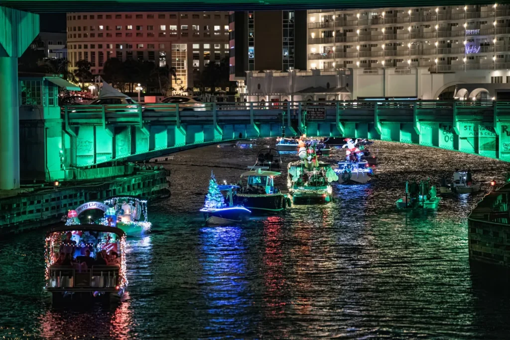 Boats decorated with festive lights and Christmas ornaments float down a river at night during a holiday boat parade.