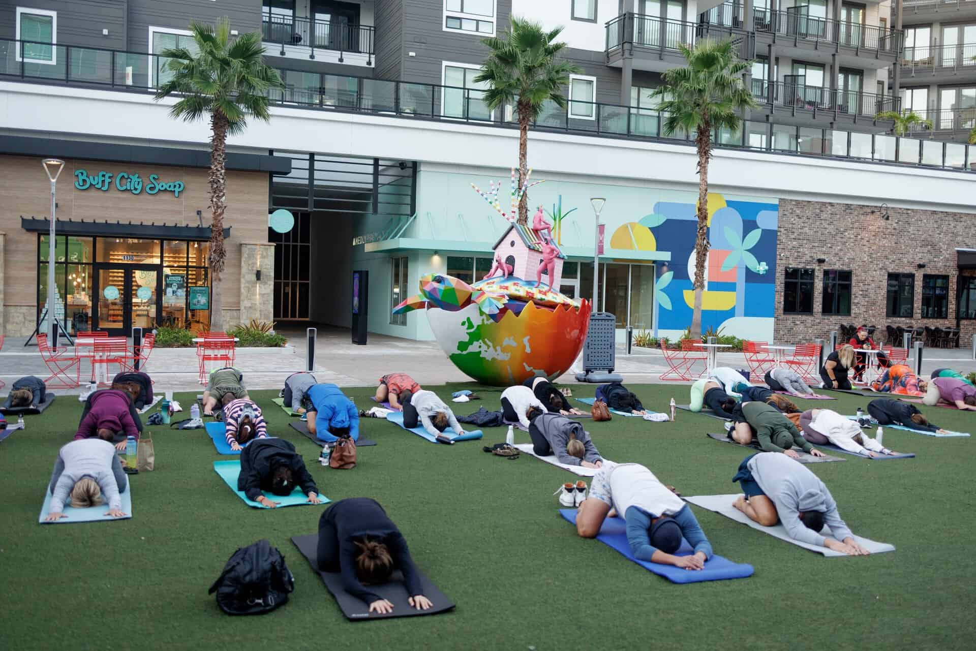 A group of people practice yoga outdoors on a lawn near a colorful sculpture and Buff City Soap storefront.