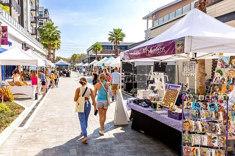 The image shows a lively outdoor market with vendor tents lining both sides of a paved walkway, featuring artwork and crafts for sale as people stroll and browse under a bright blue sky.