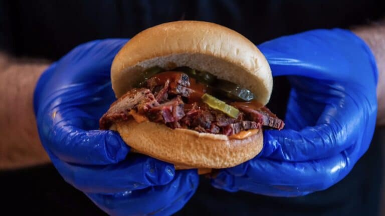 a smoked brisket sandwich held by hands in blue gloves