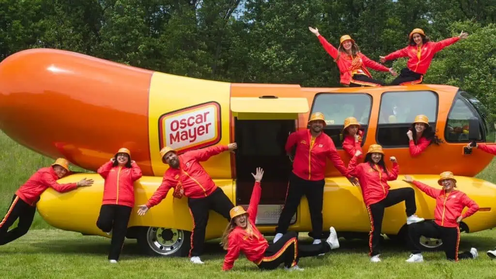 A group of people in matching red outfits pose energetically around the iconic Oscar Mayer Wienermobile.