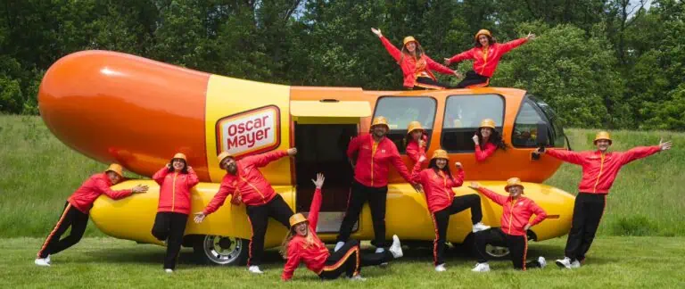 A large car built in the shape of a hot dog is parked in a field with multiple people posing in front of it in red and yellow outfits