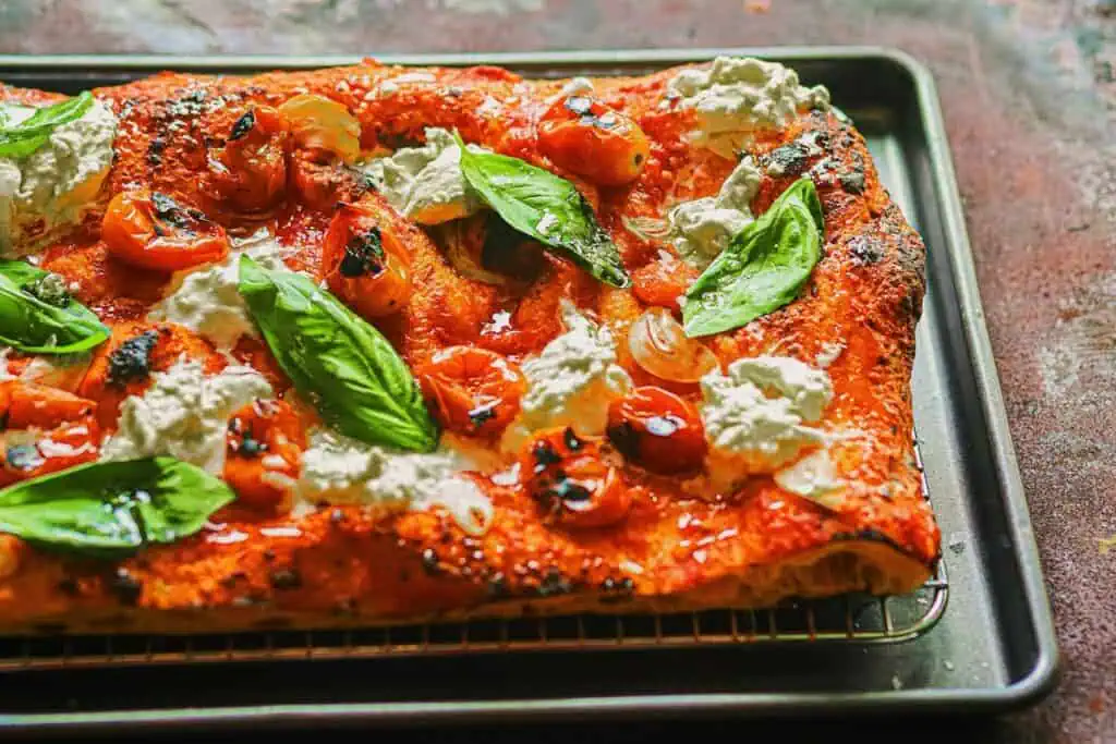 a pizza on a pan with basil leaves and fresh tomato on top. the pizza is thick and rectangular 