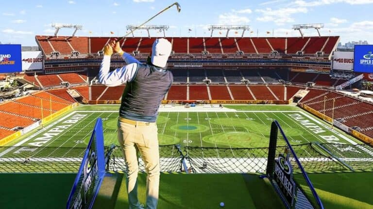 A person hits a golf ball from a tee at a large football stadium