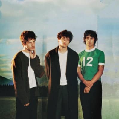 Three young men stand outdoors with a hazy sky and landscape in the background, one wearing a green jersey with the number 12.