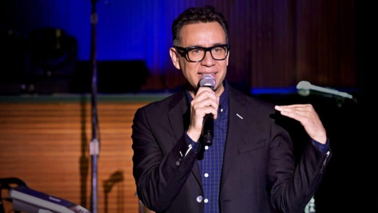 a performer on stage during a stand up comedy special. He's wearing glasses, a dark blazer and a blue button down shirt