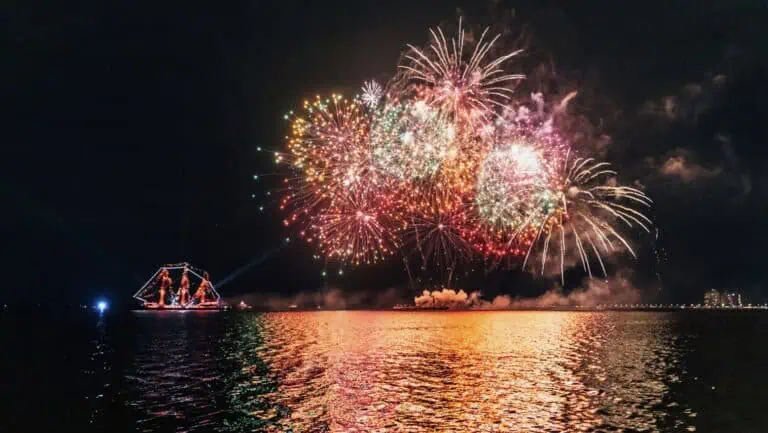 a pirate ship in the water illuminated with colorful lights while a large fireworks display occures