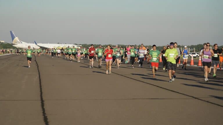 A group of people running on an airport runway. A big airplane is in the background