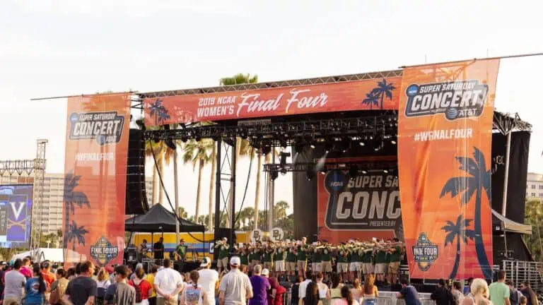 outdoor concert venue with a stage set up in a park. Orange banners frame the stage. It has branding for the NCAA Women's Final Four