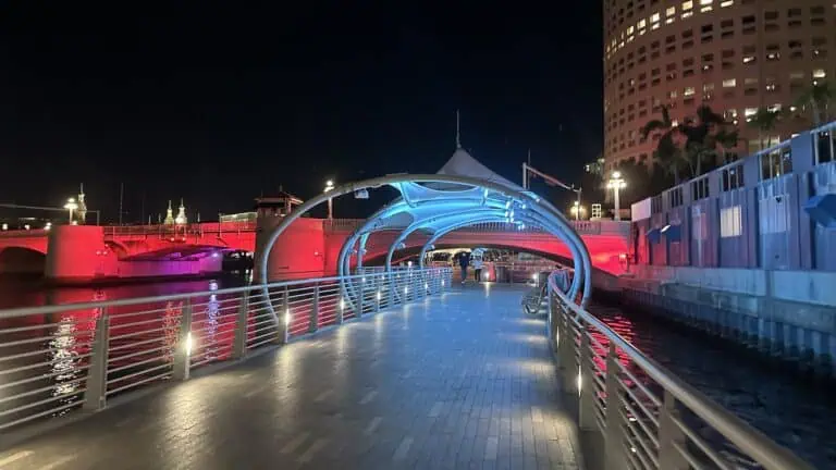 a riverwalk illuminated in red and blue lights at night. the path is crossing under a bridge